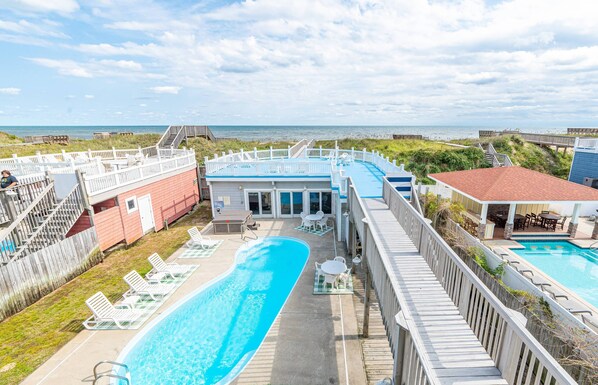 A pool area with a view of the beach, wooden deck, and a building with a blue roof.