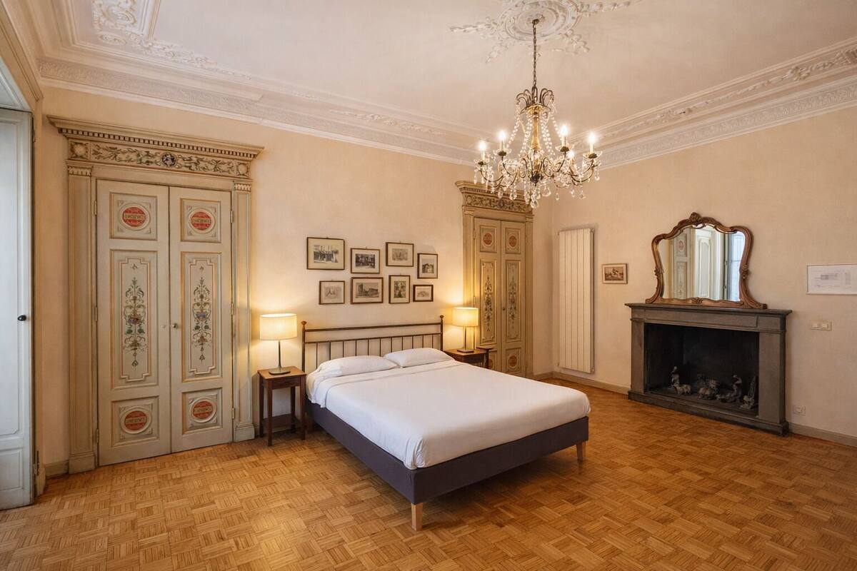 A bedroom with a large bed, two bedside tables with lamps, a chandelier, a fireplace, and a mirror above it.