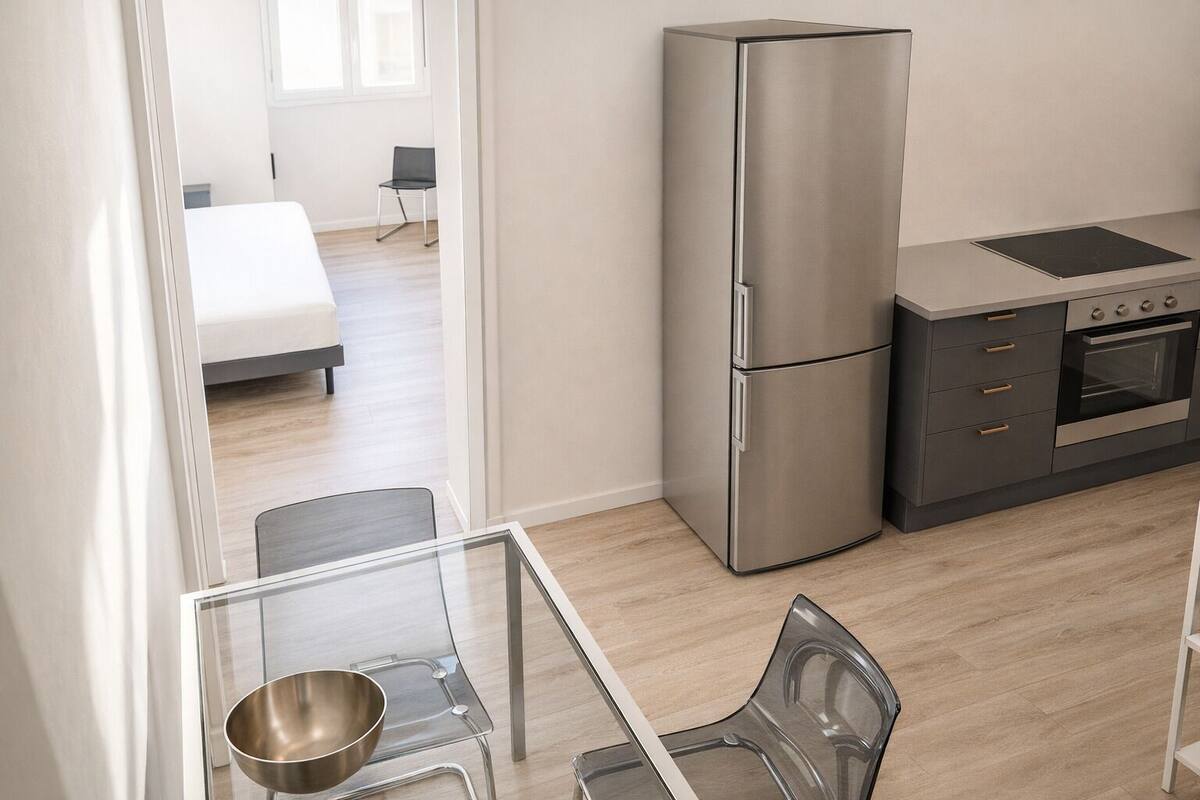 A modern kitchen with a stainless steel refrigerator, oven, and a small dining area with a glass table and chairs.