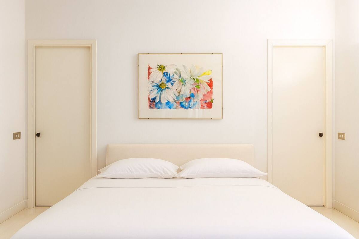 A minimalist bedroom with a large bed, two doors, and a colorful abstract painting on the wall.