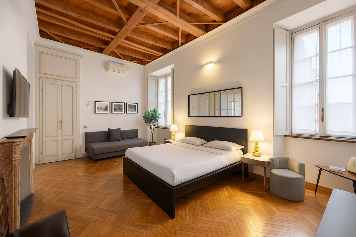 A spacious bedroom with a large bed, a wooden ceiling, a grey sofa, a small table, and a chair.