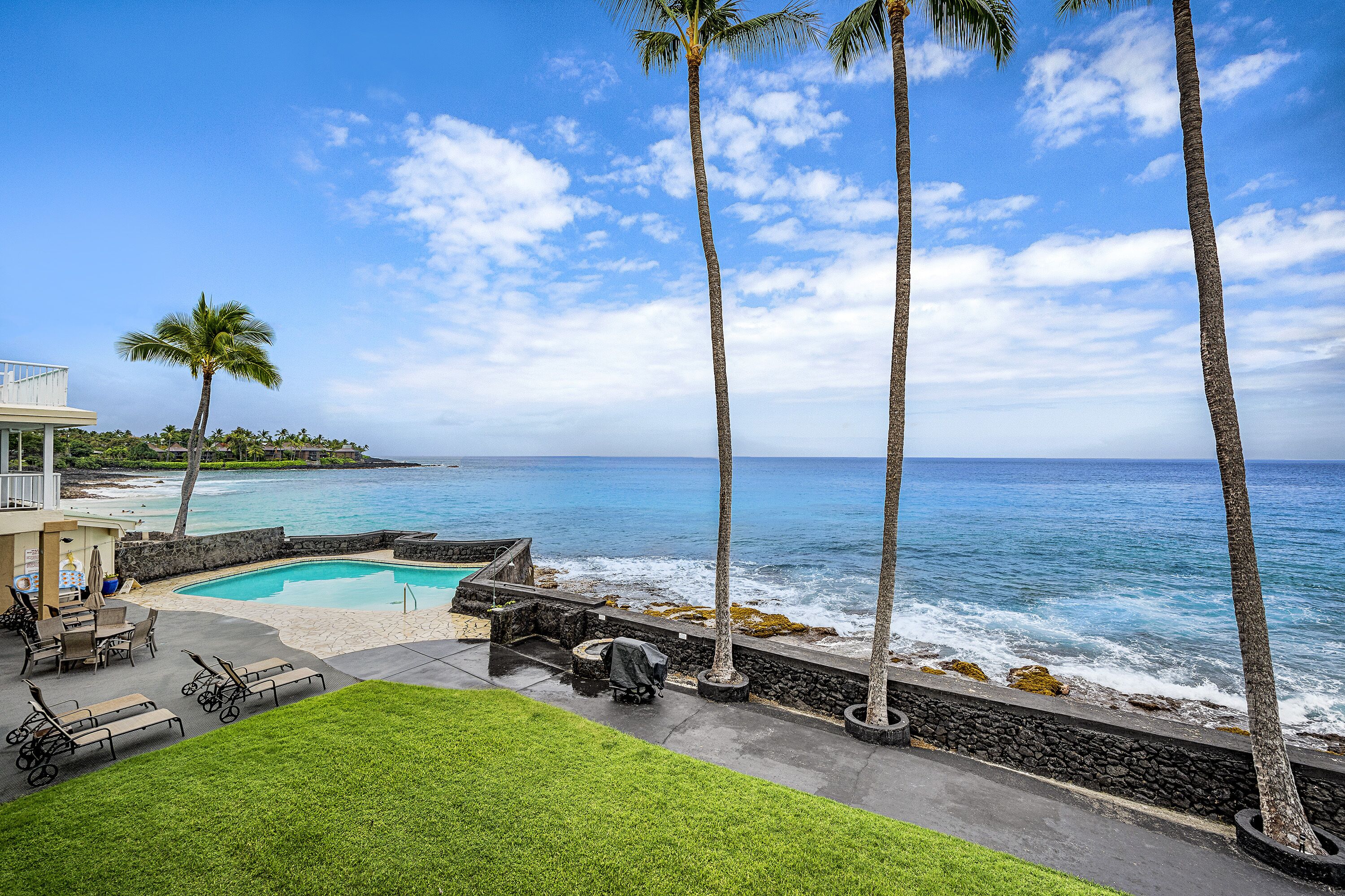 A pool area with lounge chairs, a small table, and a covered grill, overlooking the ocean with palm trees and a clear sky.