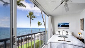 Slaapkamer met balkon en uitzicht op zee, met een bed met een boommotief, een schilderij en een plafondventilator.