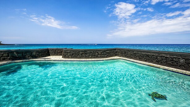 Een helderblauw zwembad met een stenen muur en uitzicht op de oceaan.