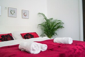 Cama bem arrumada com roupa de cama vermelha e branca, duas obras de arte em formato de coração na parede e um vaso com planta.