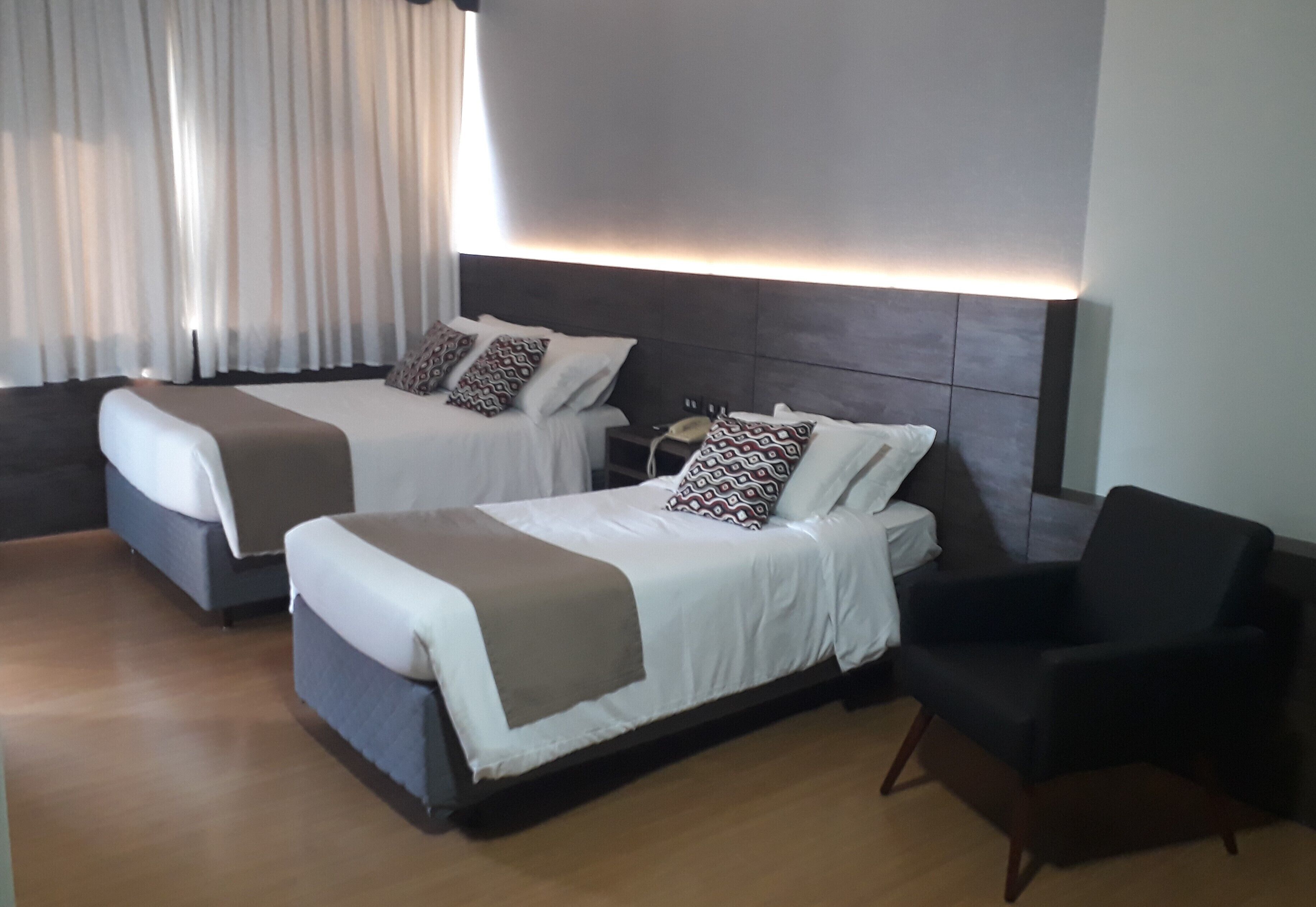 Quarto de hotel com duas camas, uma mesa de cabeceira com abajur e uma poltrona preta.