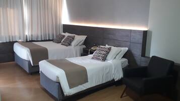 Quarto de hotel com duas camas, uma mesa de cabeceira com abajur e uma poltrona preta.