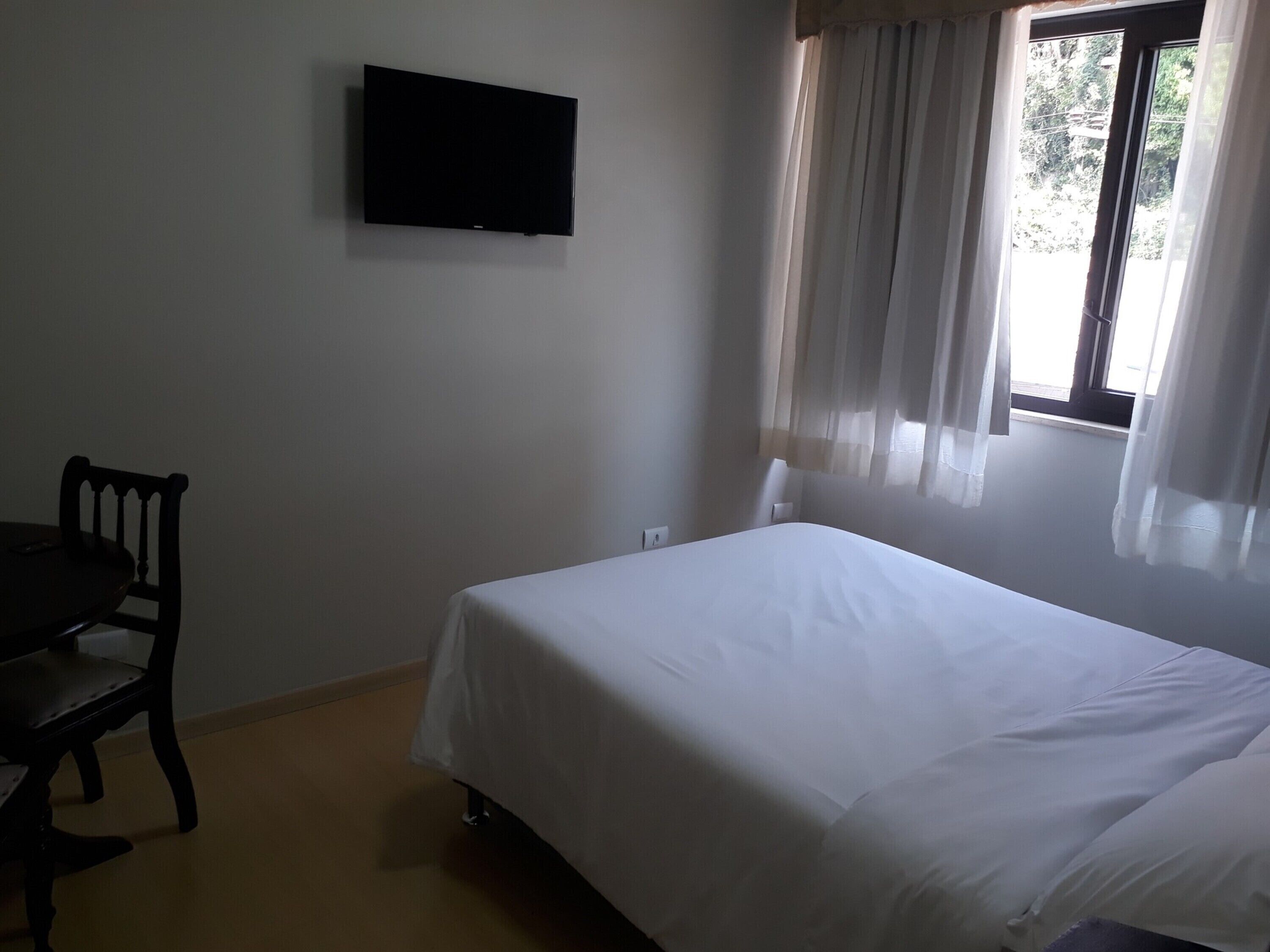 Une pièce comprenant un lit, une chaise, un téléviseur fixé au mur et une fenêtre avec des rideaux.