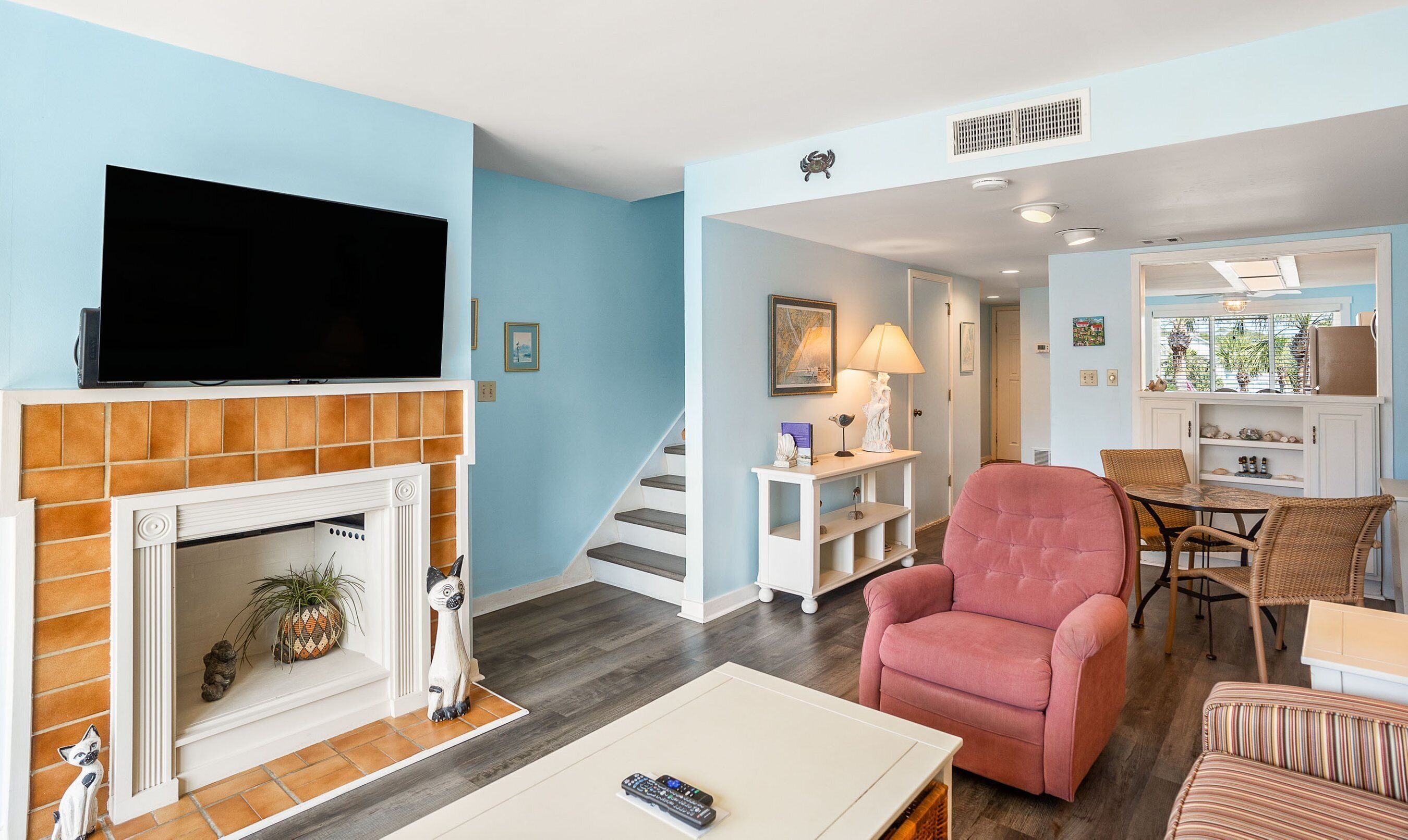 A living room with a fireplace, a television, a pink armchair, and a dining area with wicker furniture.