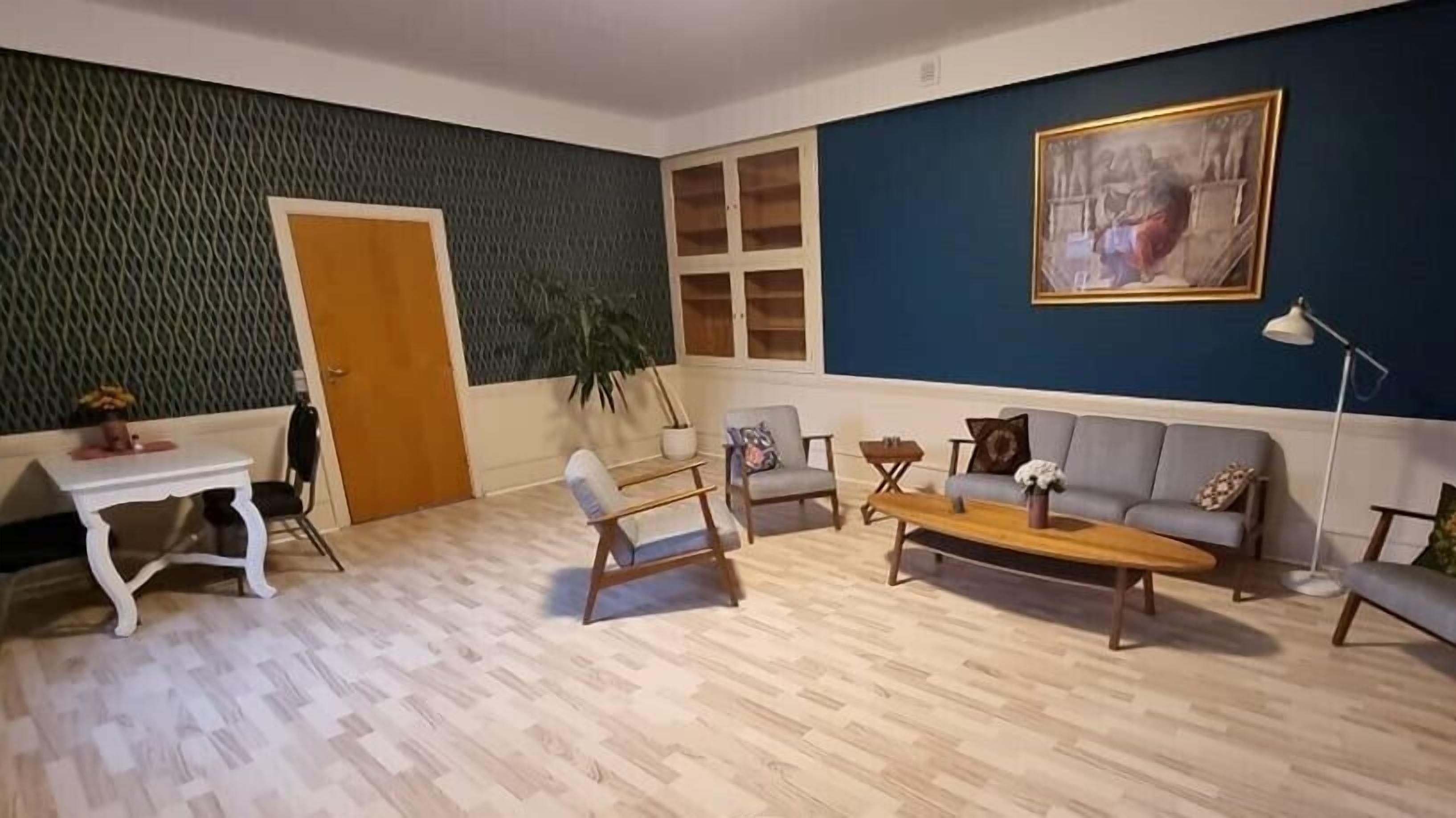 A living room with a wooden floor, a sofa, a coffee table, and a chair. There is a door, a plant, and a painting on the wall.
