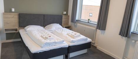 Una cama doble con sábanas y toallas blancas, una mesita de noche, cortinas en la ventana y un radiador.