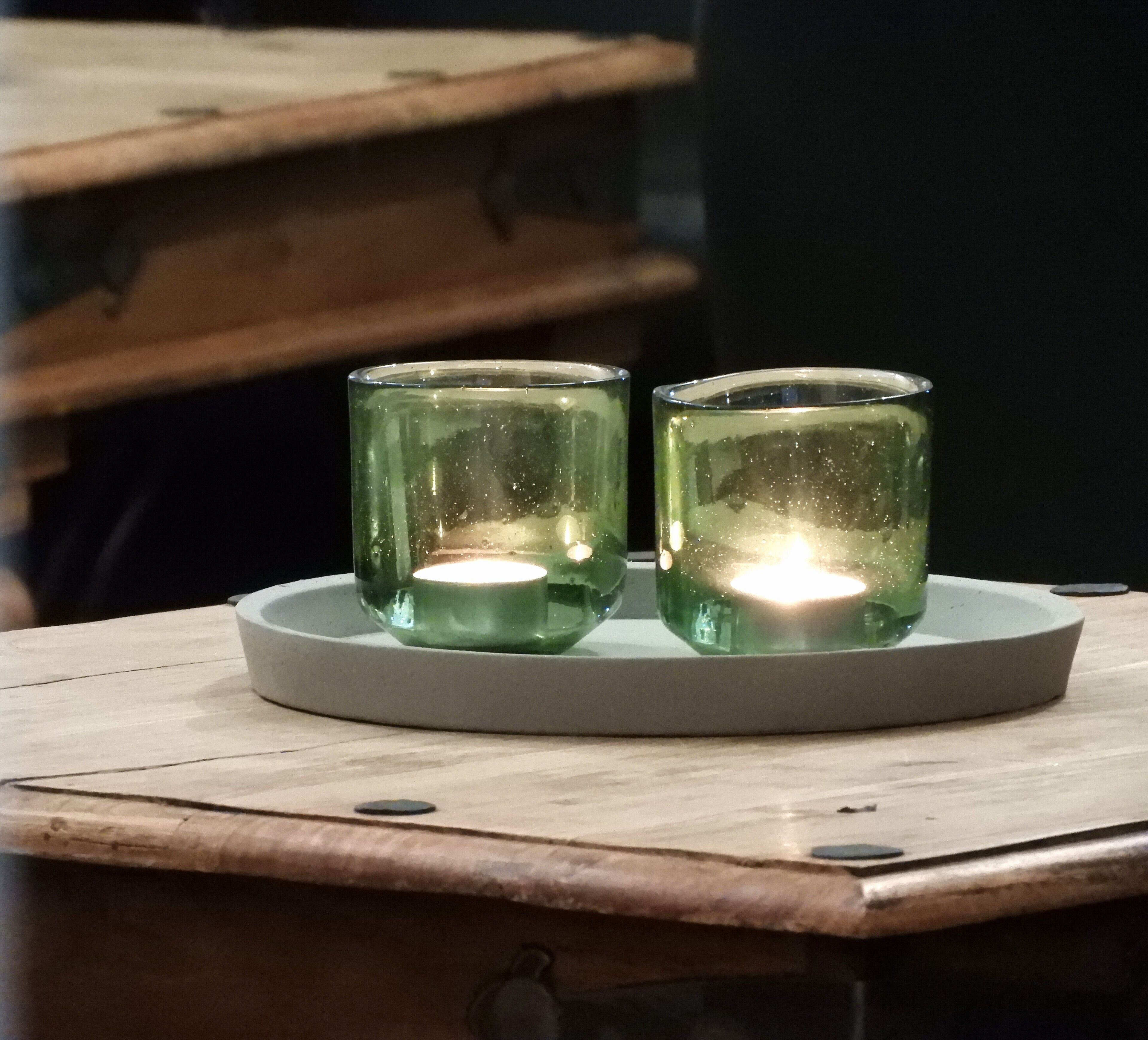 Two green glass candle holders with candles on a white tray, placed on a wooden table.