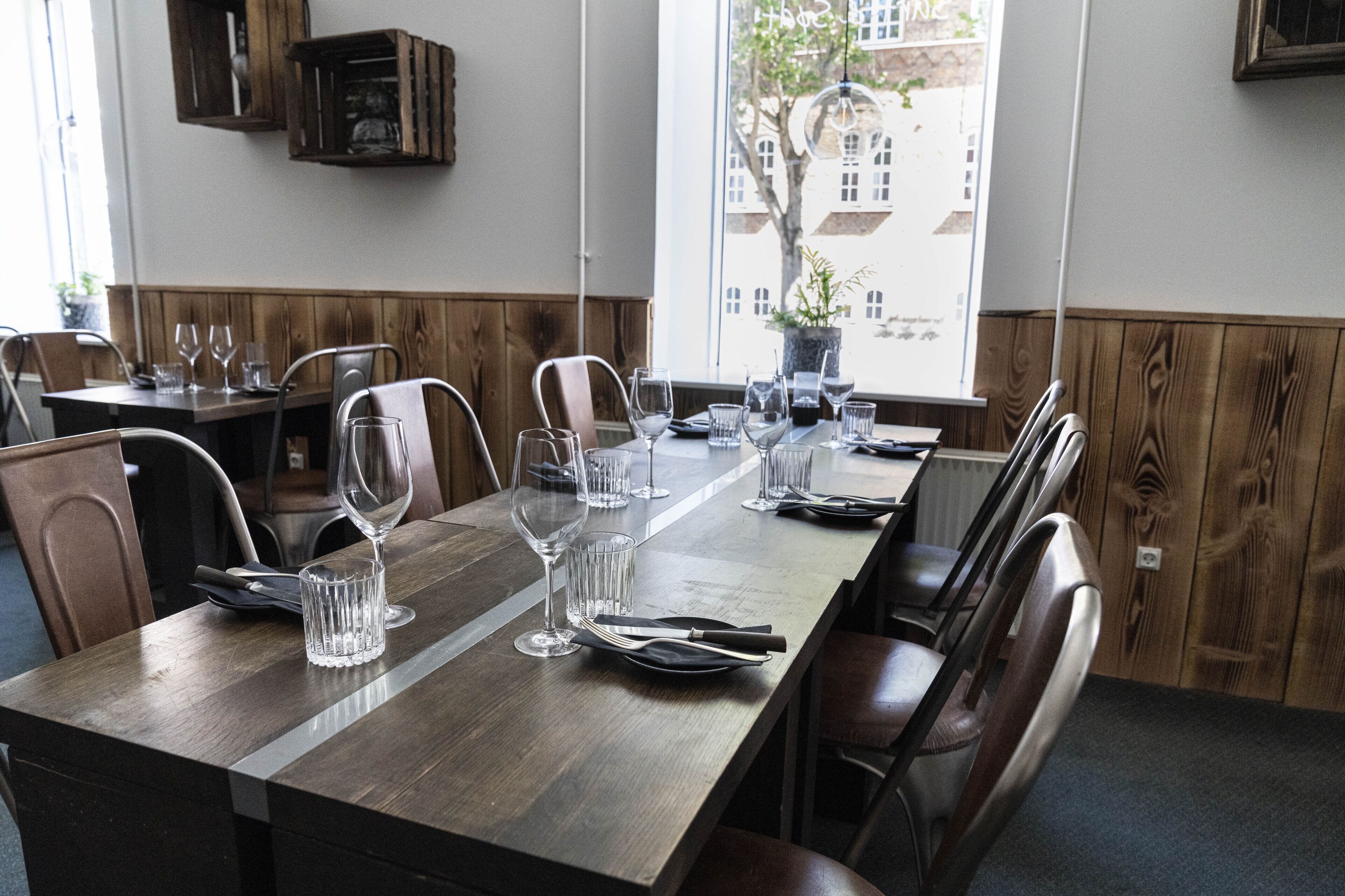 A dining area with a long wooden table set for a meal, surrounded by chairs, and a window with a view of buildings and a tree.