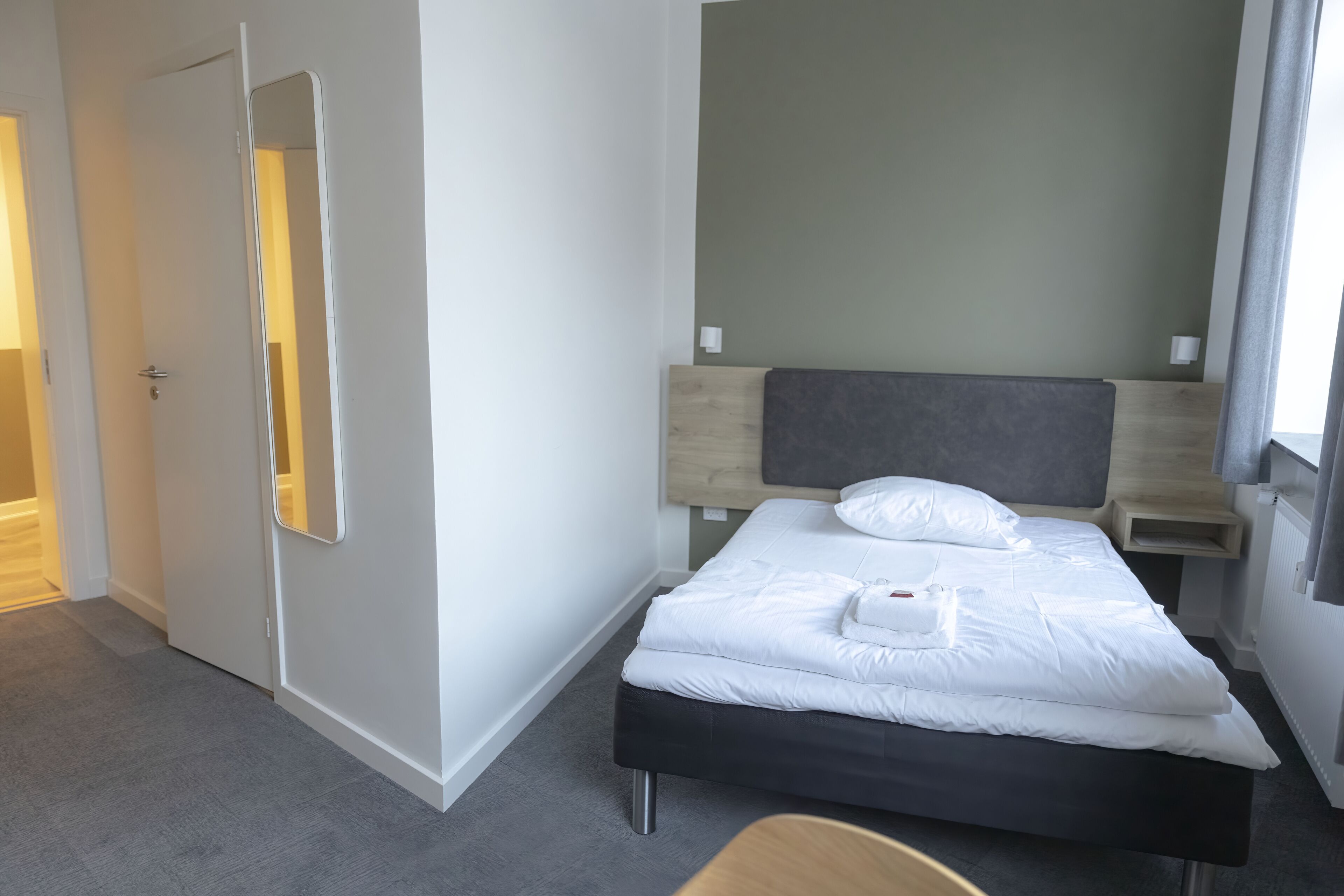 Una habitación de hotel moderna con una cama bien hecha, una mesita de noche, una silla y un espejo grande en la pared.
