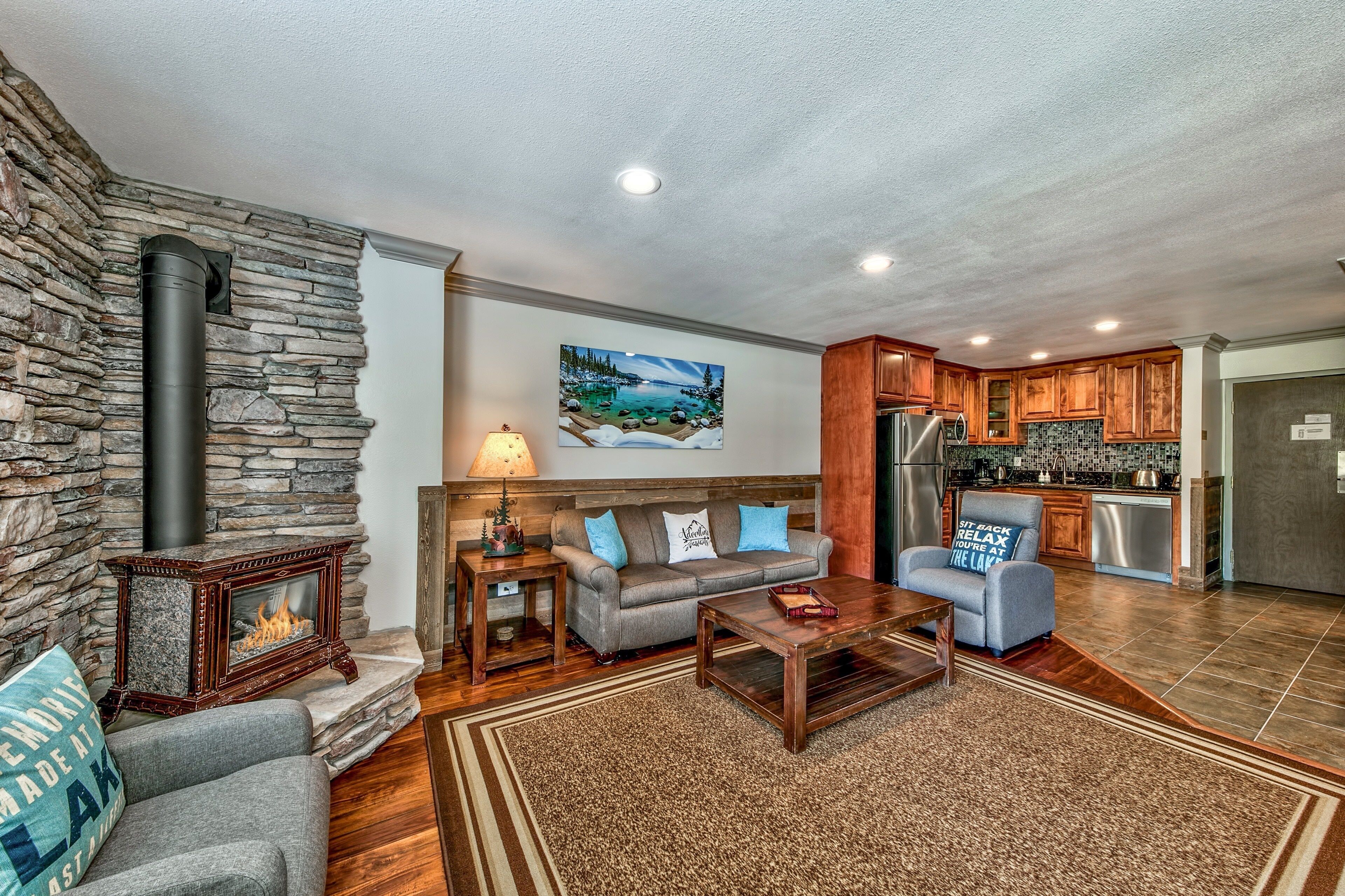 A cozy living room with a stone fireplace, a sofa, a coffee table, and a kitchen area with wooden cabinets and a stainless steel refrigerator.