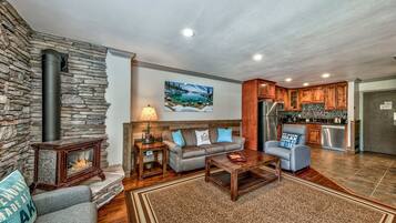 A cozy living room with a stone fireplace, a sofa, a coffee table, and a kitchen area with wooden cabinets and a stainless steel refrigerator.