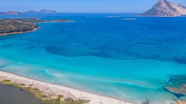 An aerial view of a coastal area with clear turquoise waters, a sandy beach, and rocky outcrops in the distance.