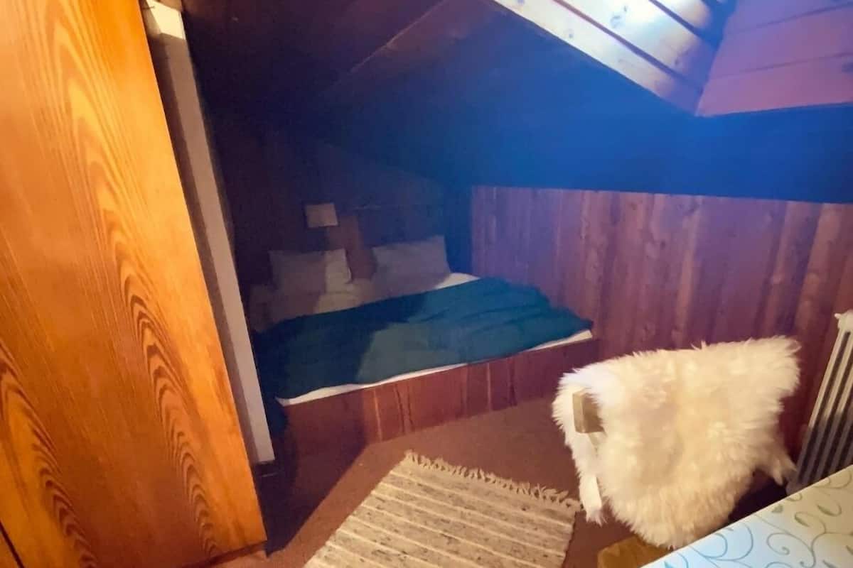 Une petite pièce chaleureuse aux murs en bois et au plafond incliné. On aperçoit un lit recouvert d’une couverture verte, et un chien blanc et duveteux est assis sur une chaise.