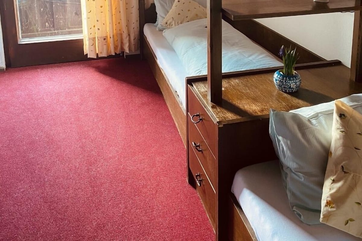 Une chambre avec un lit superposé, une petite table agrémentée d’un objet décoratif, une fenêtre avec des rideaux et un tapis rouge.