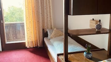 Una habitació amb una llitera, una taula petita amb un objecte decoratiu, una finestra amb cortines i una catifa vermella.