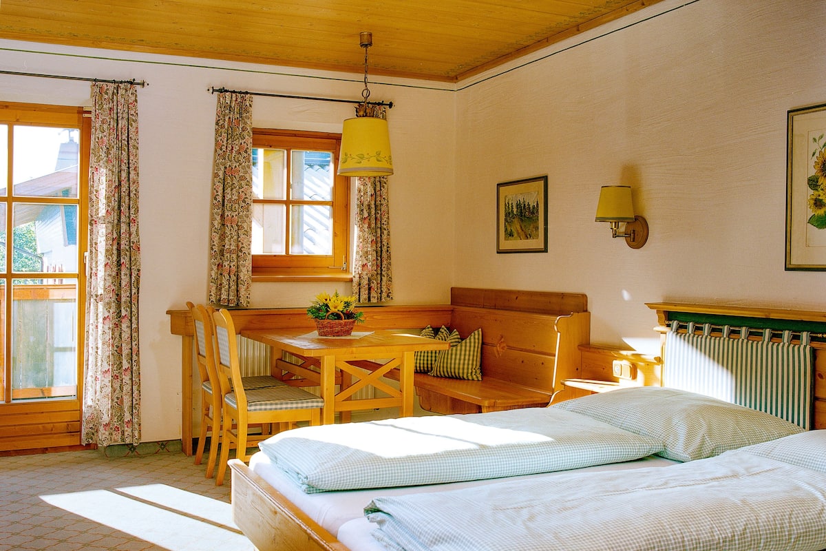 Une pièce confortable avec un lit, une table en bois entourée de chaises, un banc et une fenêtre avec des rideaux.