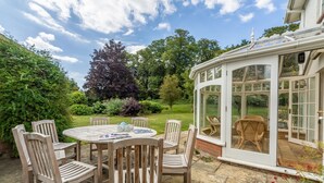 A patio with a round wooden table and chairs, a conservatory with glass doors, and a garden with various plants and trees. - Beacon Lodge, Burnham Market, Norfolk (Burnham Market)