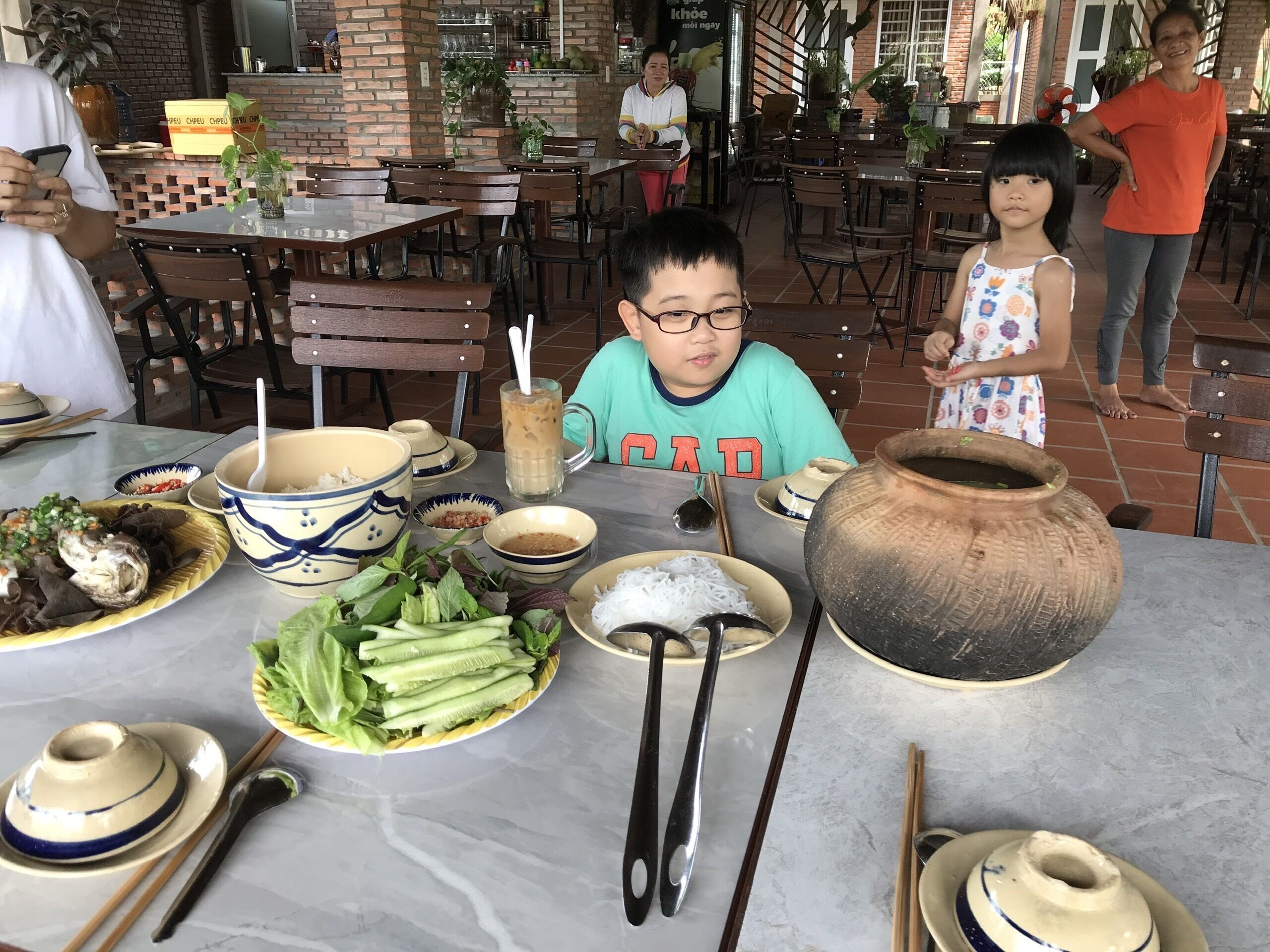 Ambiente de restaurante com mesa posta para refeição, incluindo tigelas, hashis e um grande pote de barro.