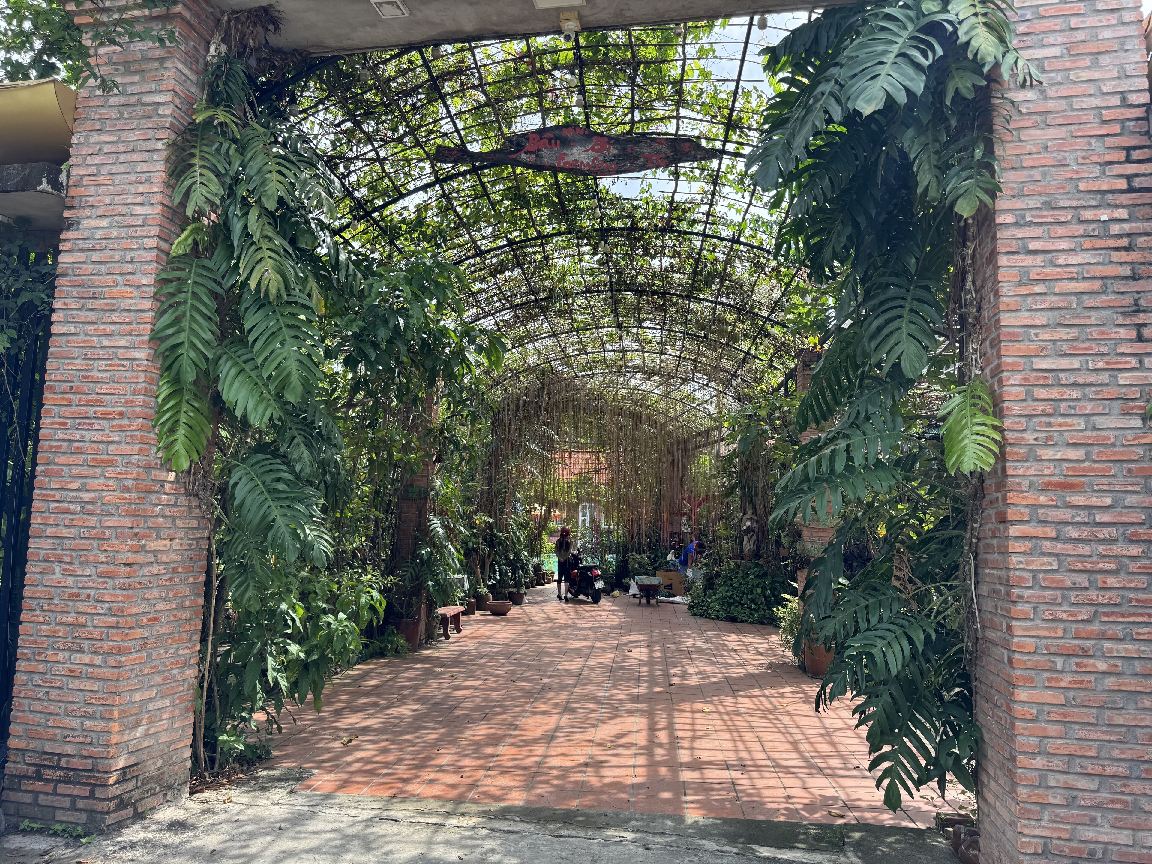 Um arco de tijolos que dá acesso a um jardim coberto, com plantas em vasos e um banco.