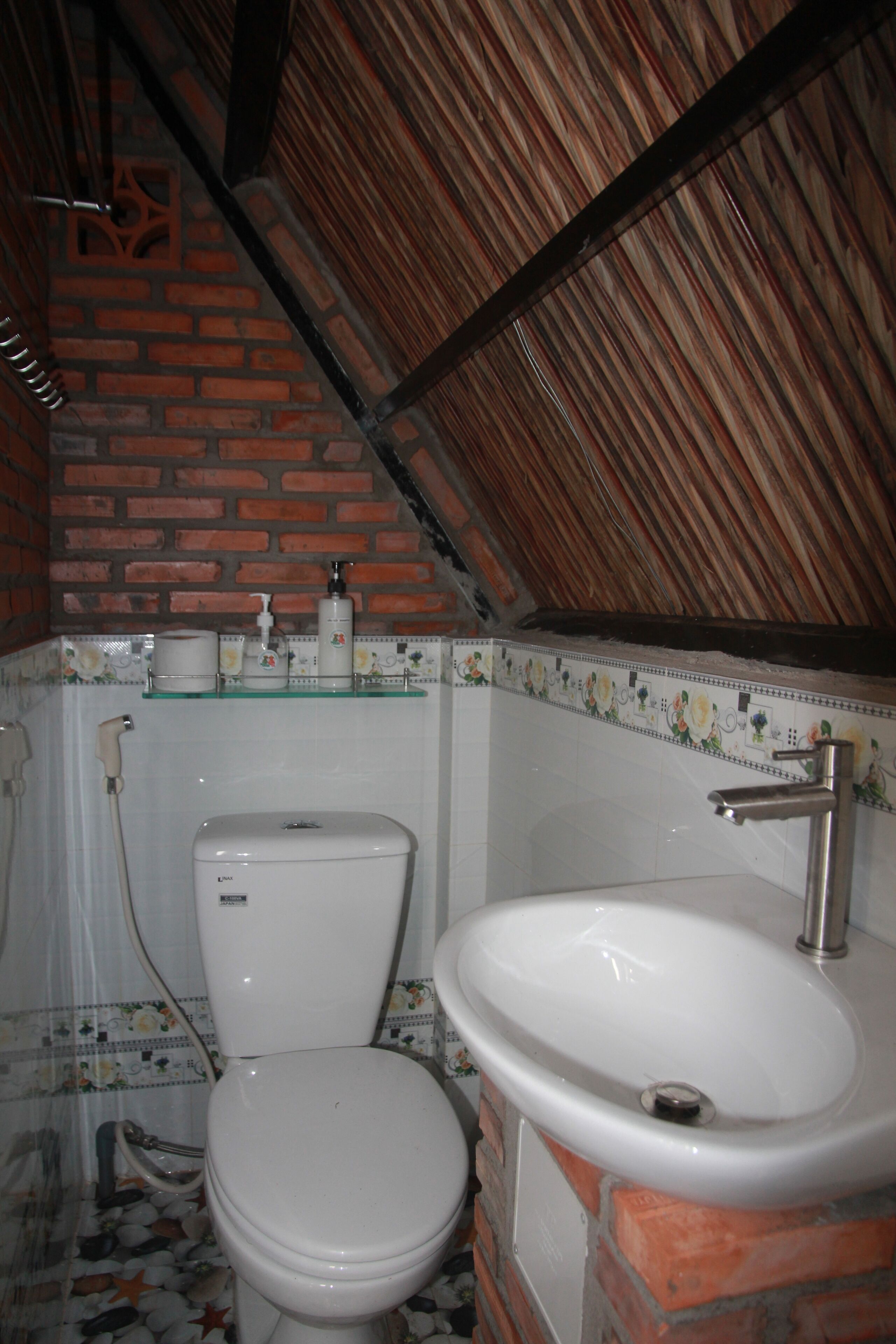 Un baño con inodoro, lavamanos y ducha. Las paredes están cubiertas de azulejos con un estampado floral, y hay una pared de acento de ladrillo.