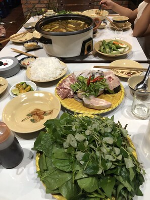 A table set with various dishes including a large pot of soup, rice, meat, vegetables, and a bowl of greens. - Bau Bi Mini Farmstay Mekong - Tien Giang (Trung An)