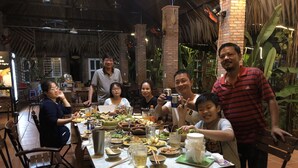 Un grupo de personas cenando en un restaurante con techo de paja y paredes de ladrillo, mostrando una mesa larga con diversos platos y bebidas.