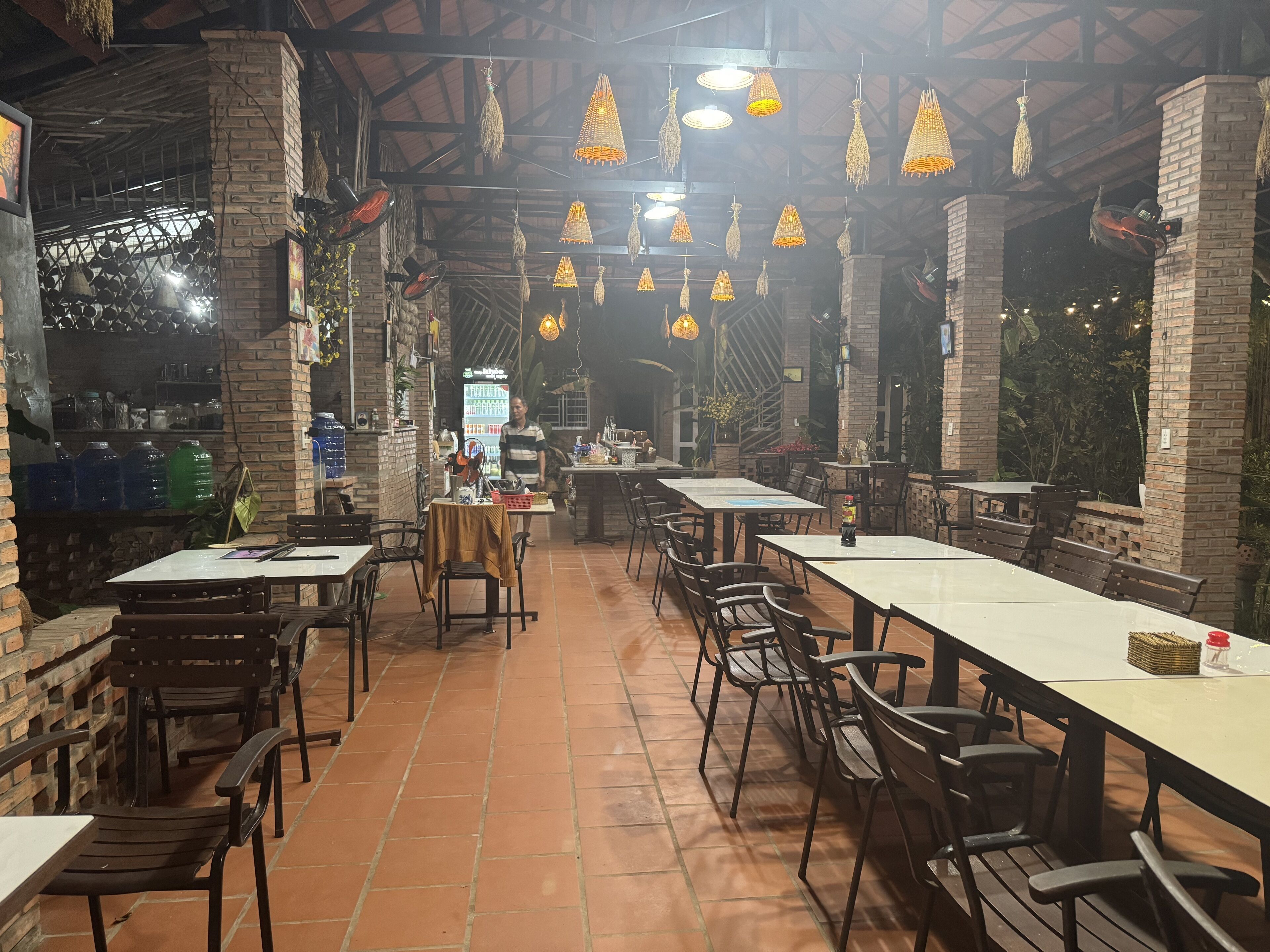 Un restaurant situé à l’intérieur, avec des murs en briques, des luminaires suspendus et un sol carrelé. Des tables et des chaises sont disposées pour accueillir les clients.