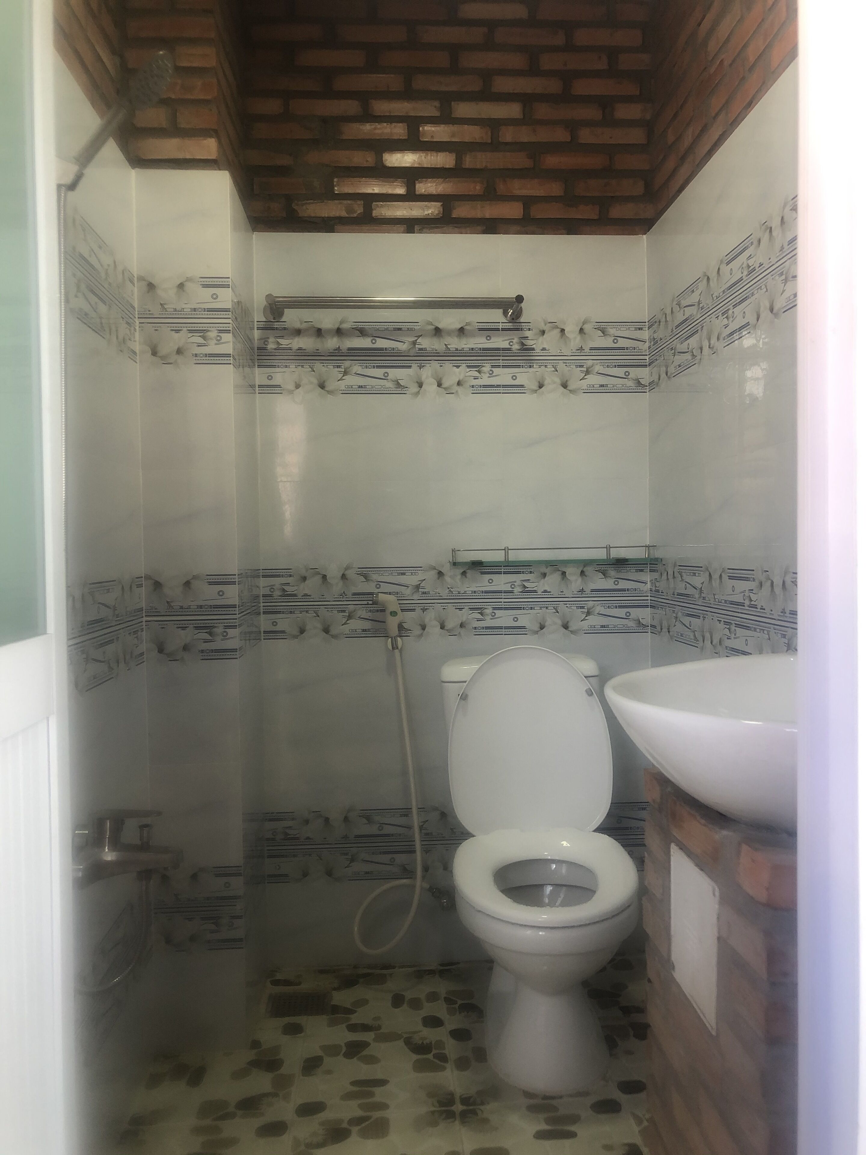 Un baño pequeño con inodoro, ducha y lavamanos. Las paredes tienen un diseño estampado y el suelo, baldosas con diseño moteado.