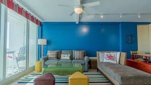 A living room with a blue wall, a ceiling fan, a sofa, a coffee table, and a window overlooking a balcony with chairs.