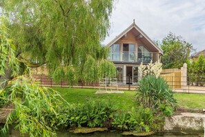 Une maison en bois à deux étages, dotée d’un balcon, entourée d’une végétation luxuriante et d’un petit étang.