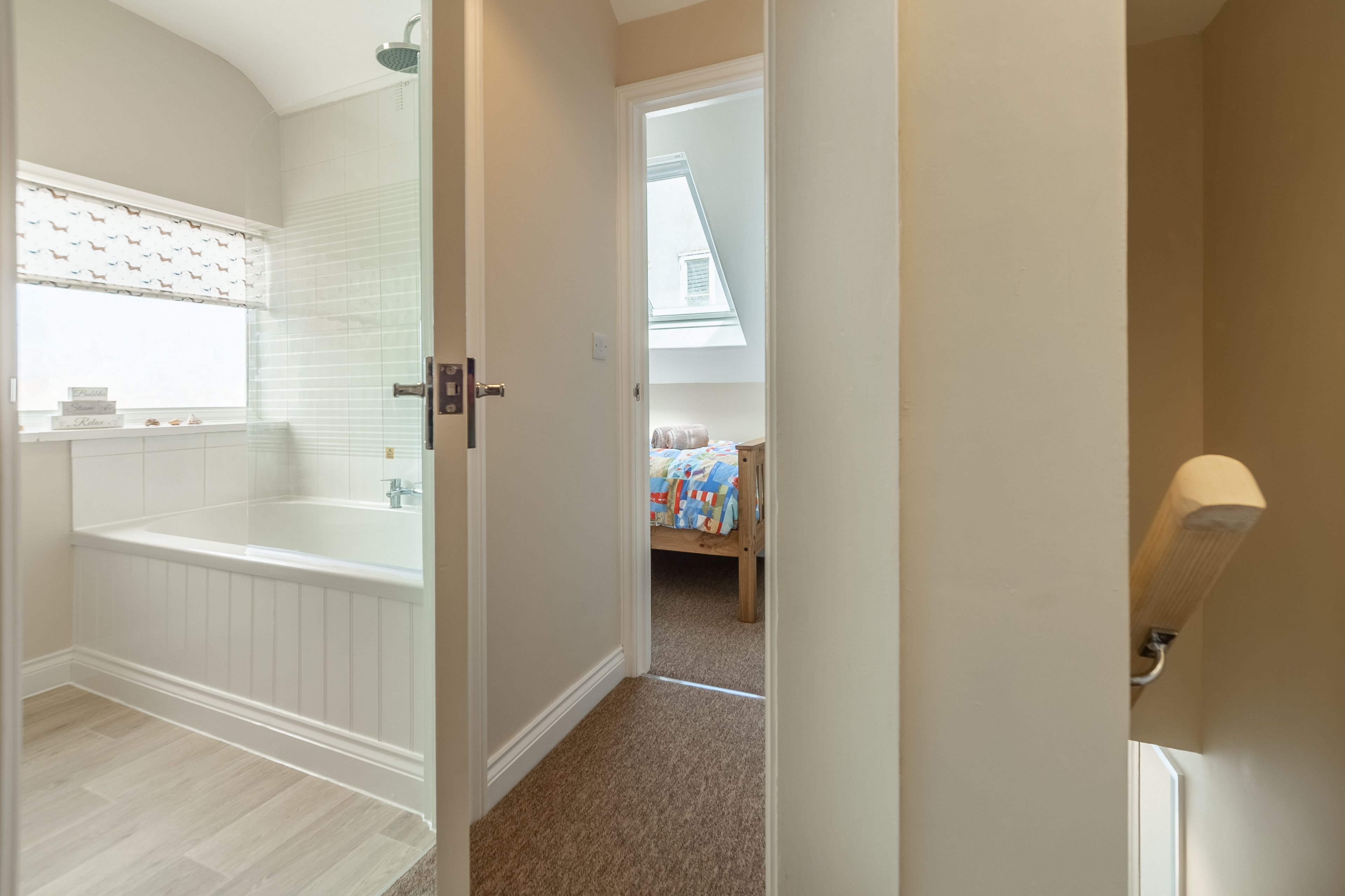 A bathroom with a bathtub, a window with patterned blinds, and a visible bedroom with a bed and a nightstand.