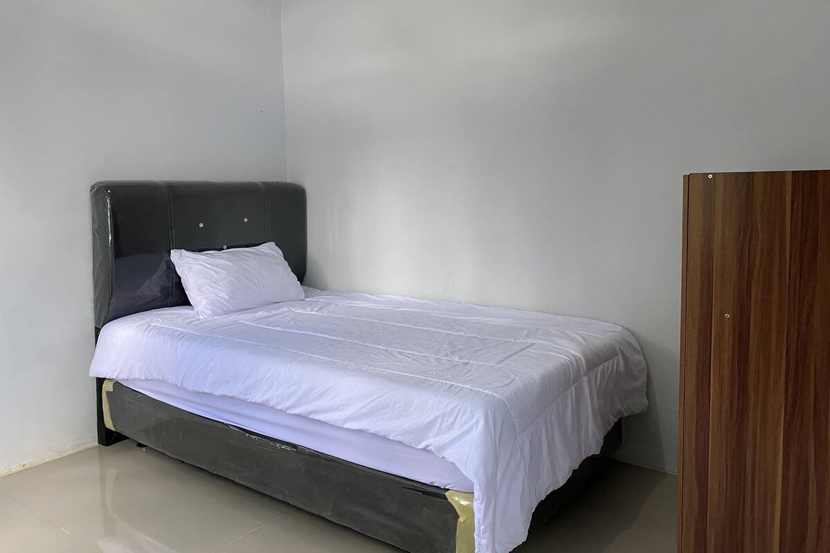Een eenpersoonsbed met een donker hoofdbord en witte beddengoed in een kamer met effen muren en een houten garderobe.
