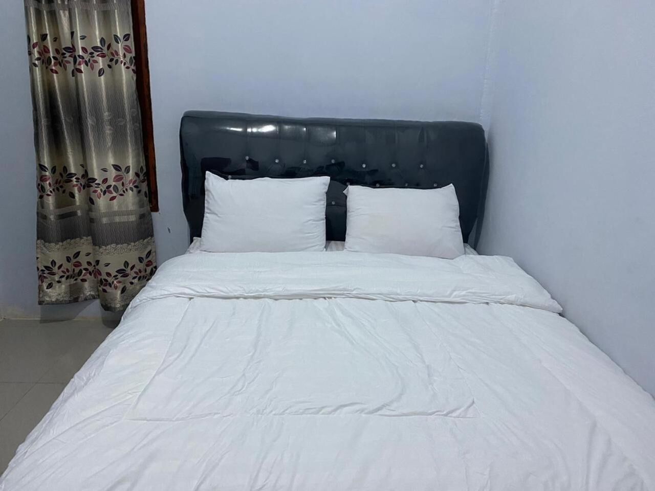 Een bed met een donker hoofdbord en witte beddengoed in een kamer met lichtgekleurde muren en een bloemen gordijn.