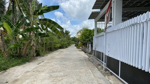 Uma estrada estreita e sem asfalto, ladeada por vegetação densa e uma cerca de metal branco.