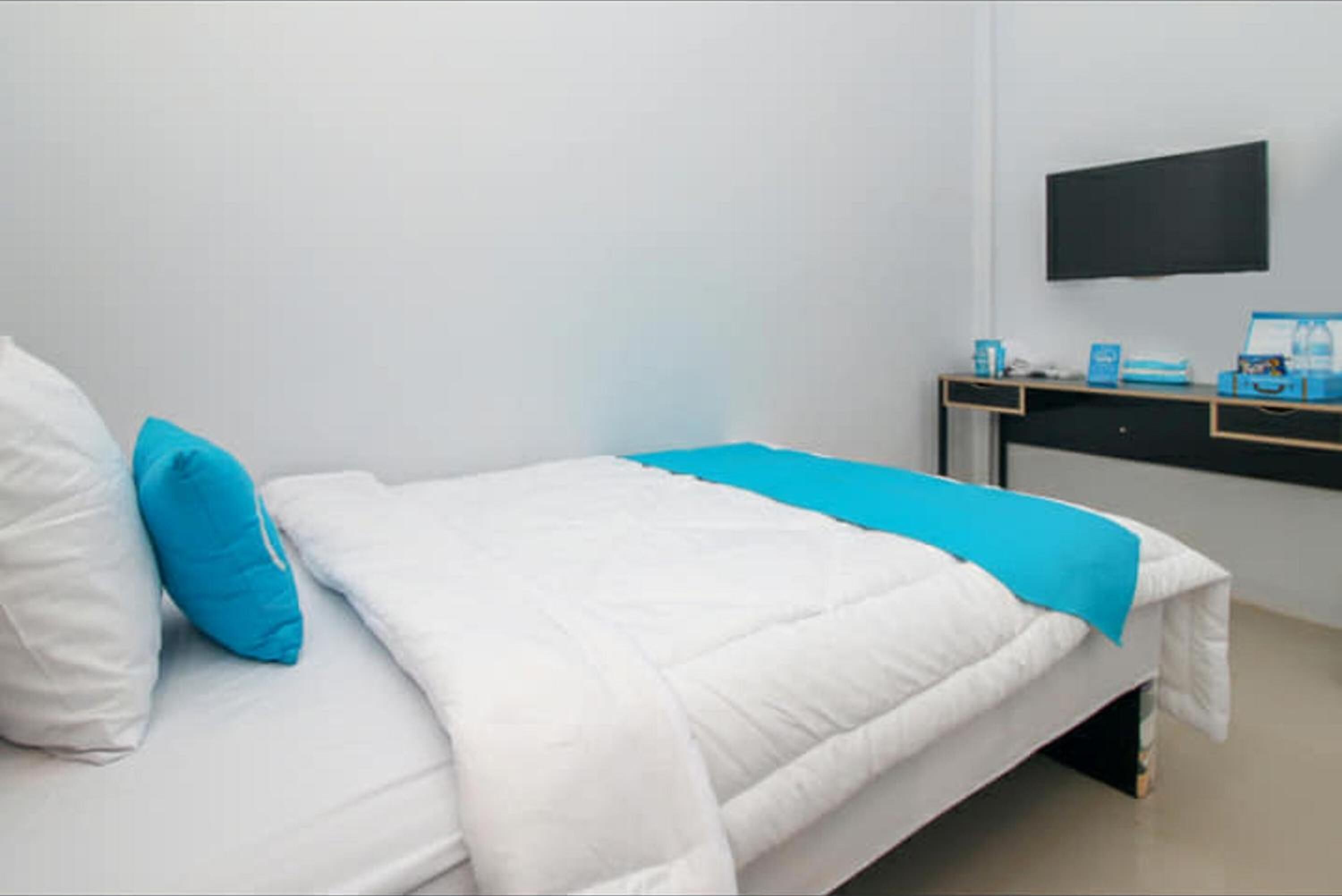 Un lit bien fait, avec une literie blanche et bleue, un téléviseur noir fixé au mur et un bureau sur lequel se trouvent un ordinateur et quelques objets.