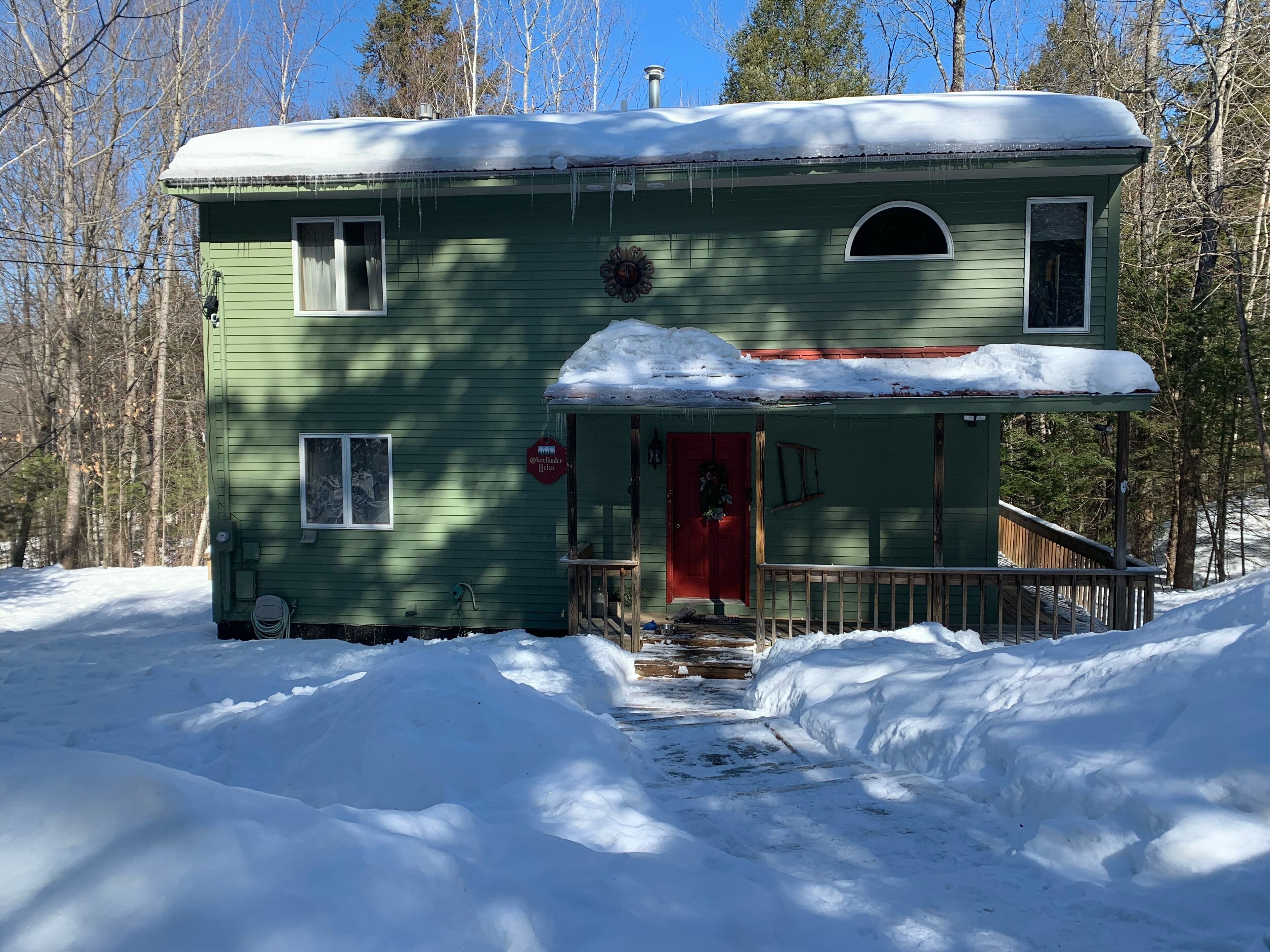 A green house with a red door, surrounded by snow, with a porch and a chimney.