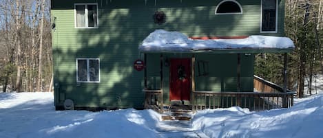 A green house with a red door, surrounded by snow, with a porch and a chimney.