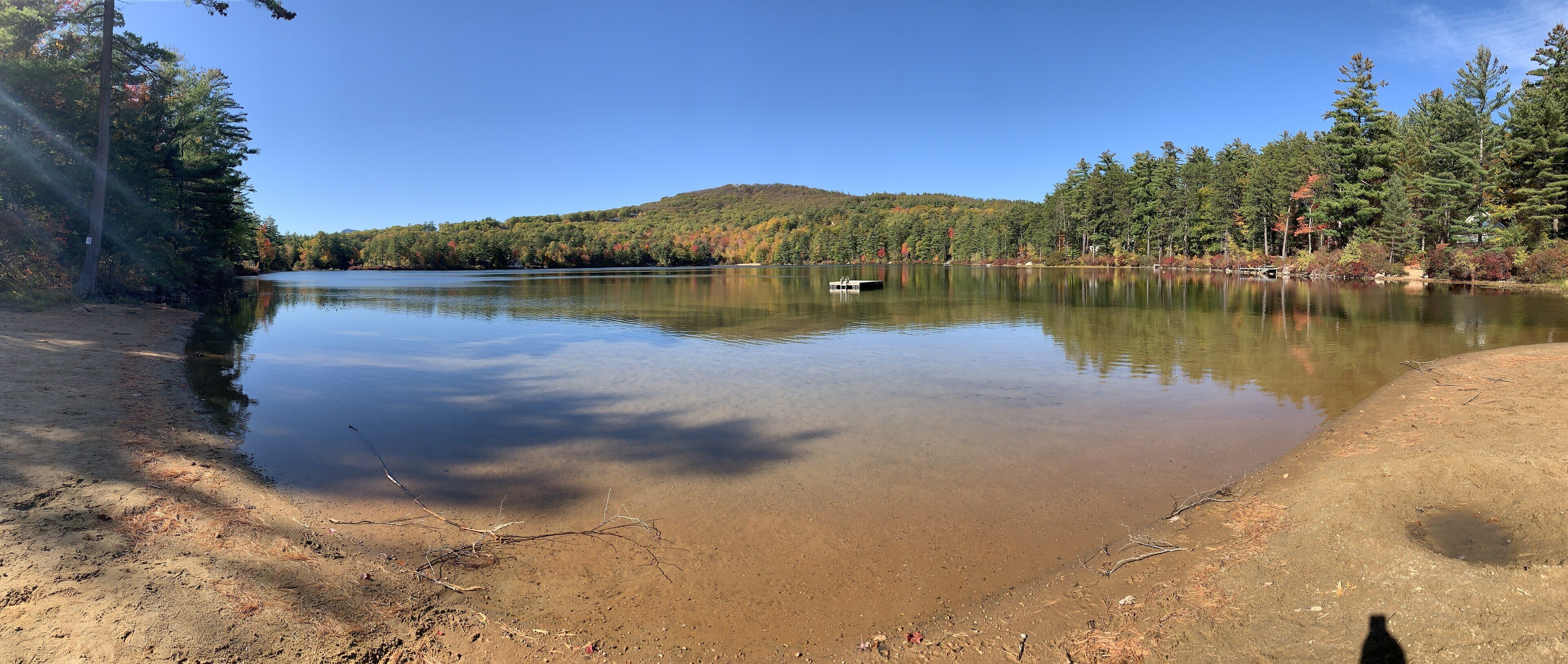 Un lago sereno con un muelle, rodeado de un bosque denso y una colina al fondo.