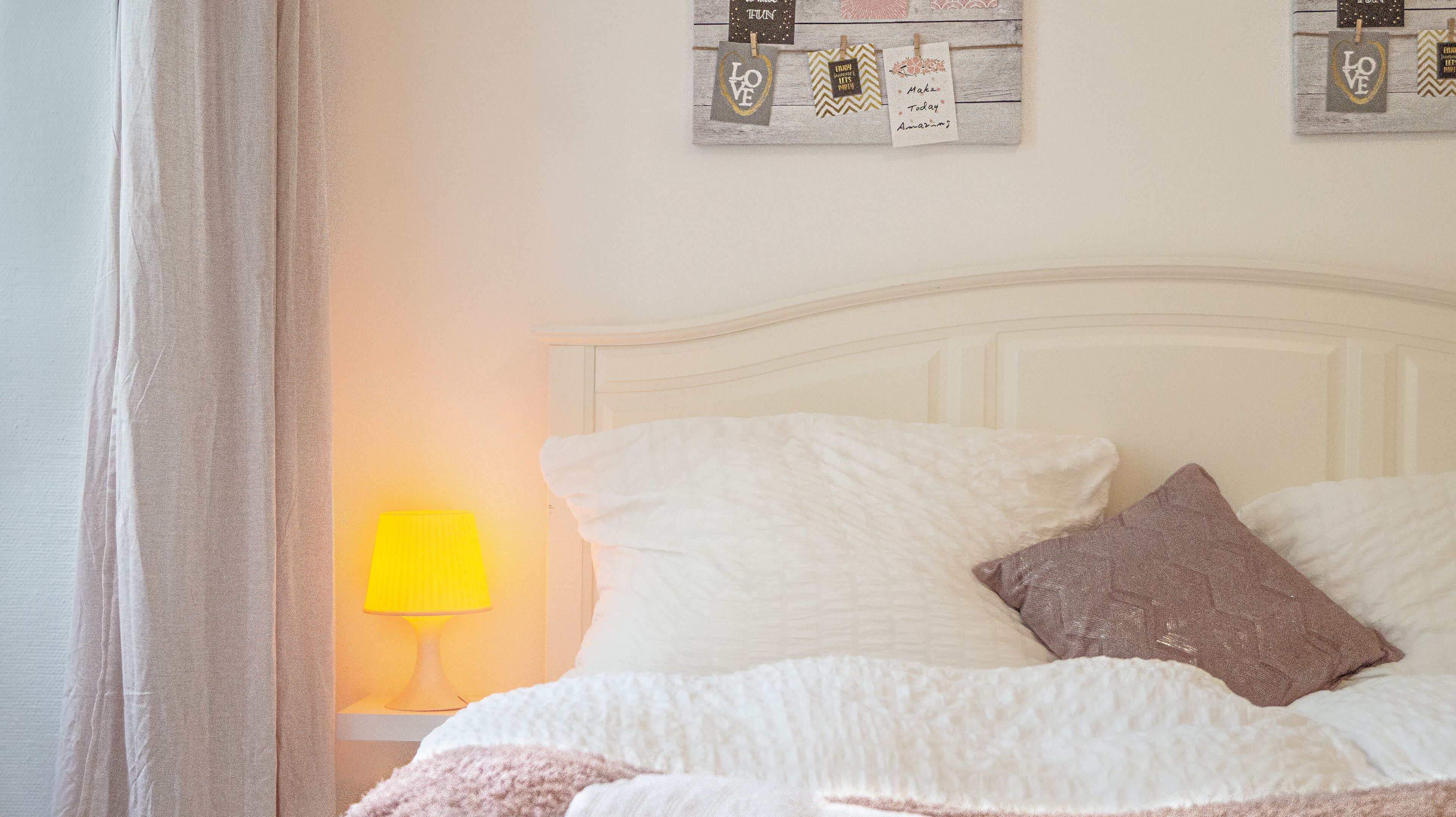 A bed with white bedding and a brown pillow, a bedside lamp, and a wall with decorative items.
