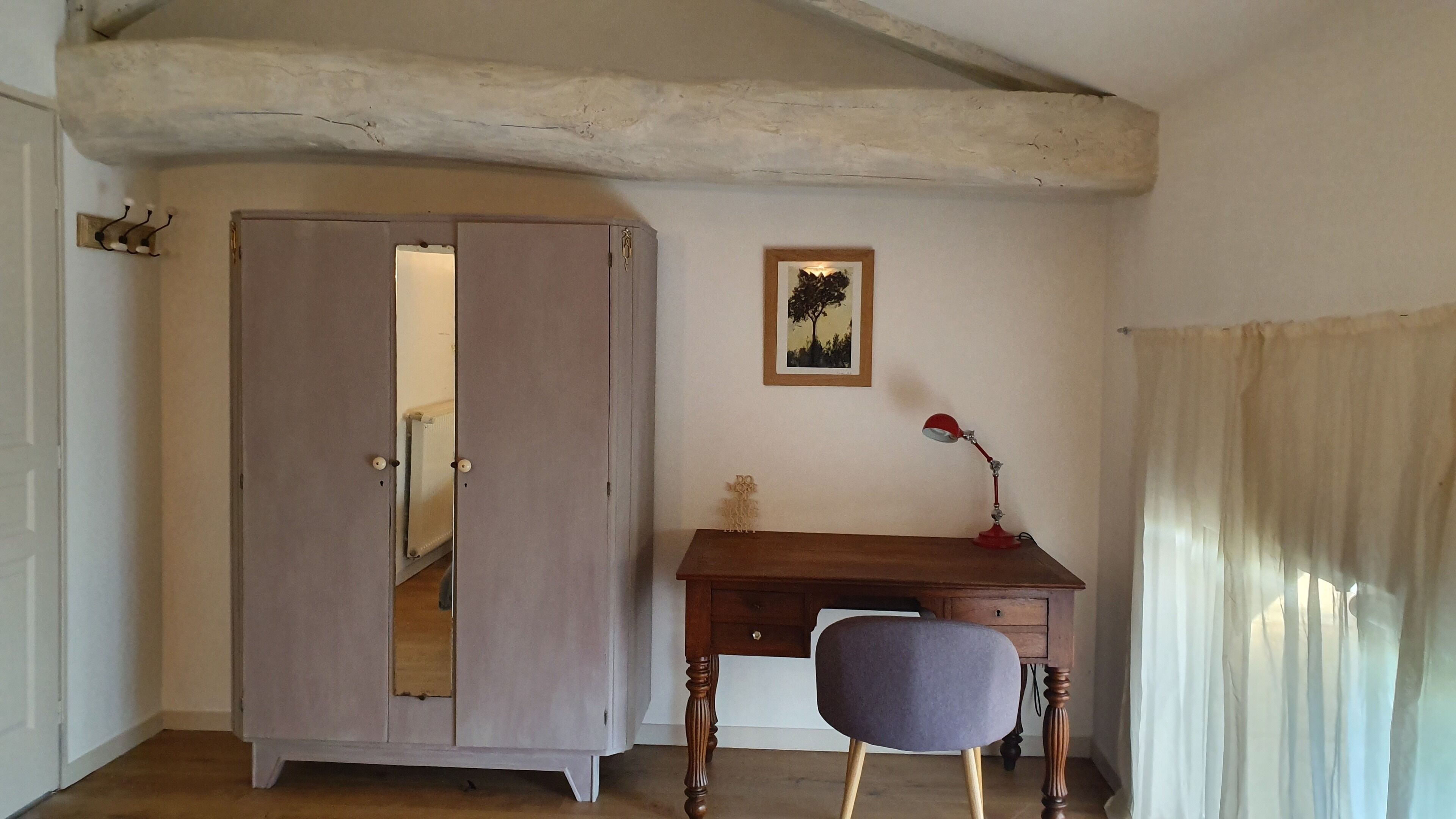 Una stanza con una scrivania in legno, una sedia, un armadio, un quadro appeso al muro e una finestra con tende.