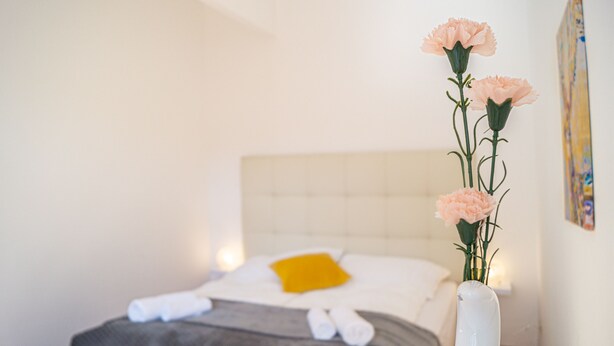 A neatly made bed with a white duvet, a yellow pillow, and a white vase with pink flowers. - Shared Modern Apartment Schönbrunn - Budget Chic Room (Vienna)