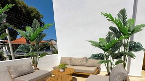 A sunlit outdoor seating area with a wooden table, cushioned chairs, and large potted plants. - Monk Island Guesthouse (Foça)
