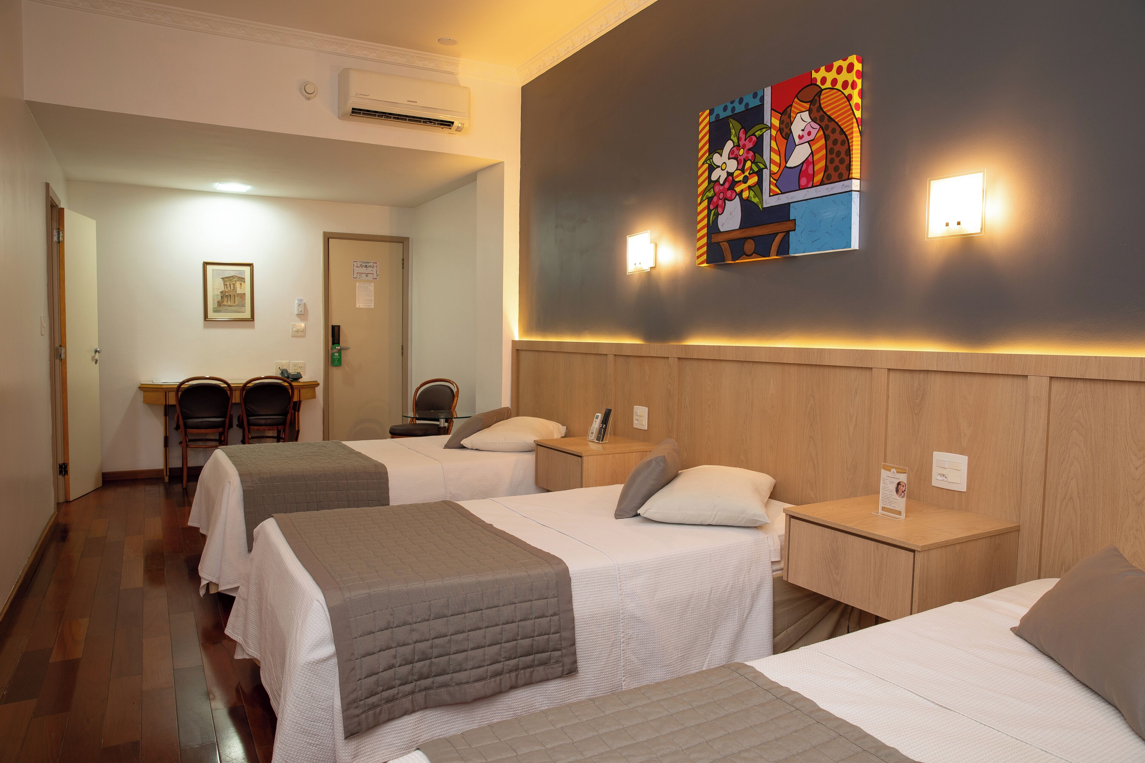 Une chambre d’hôtel avec trois lits, un bureau, des chaises et un tableau au mur.