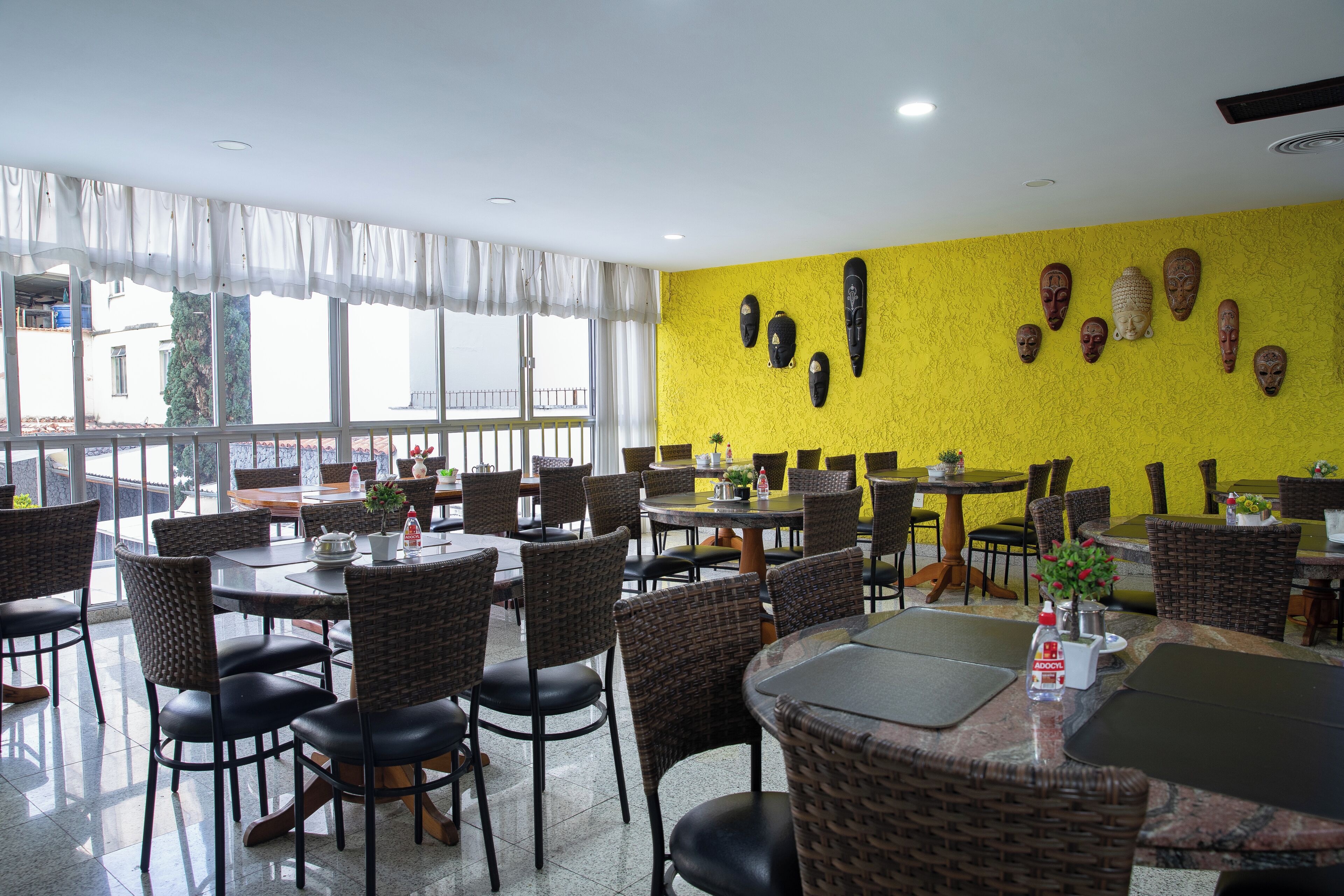A restaurant with wicker chairs and tables, a yellow wall with masks, and large windows.