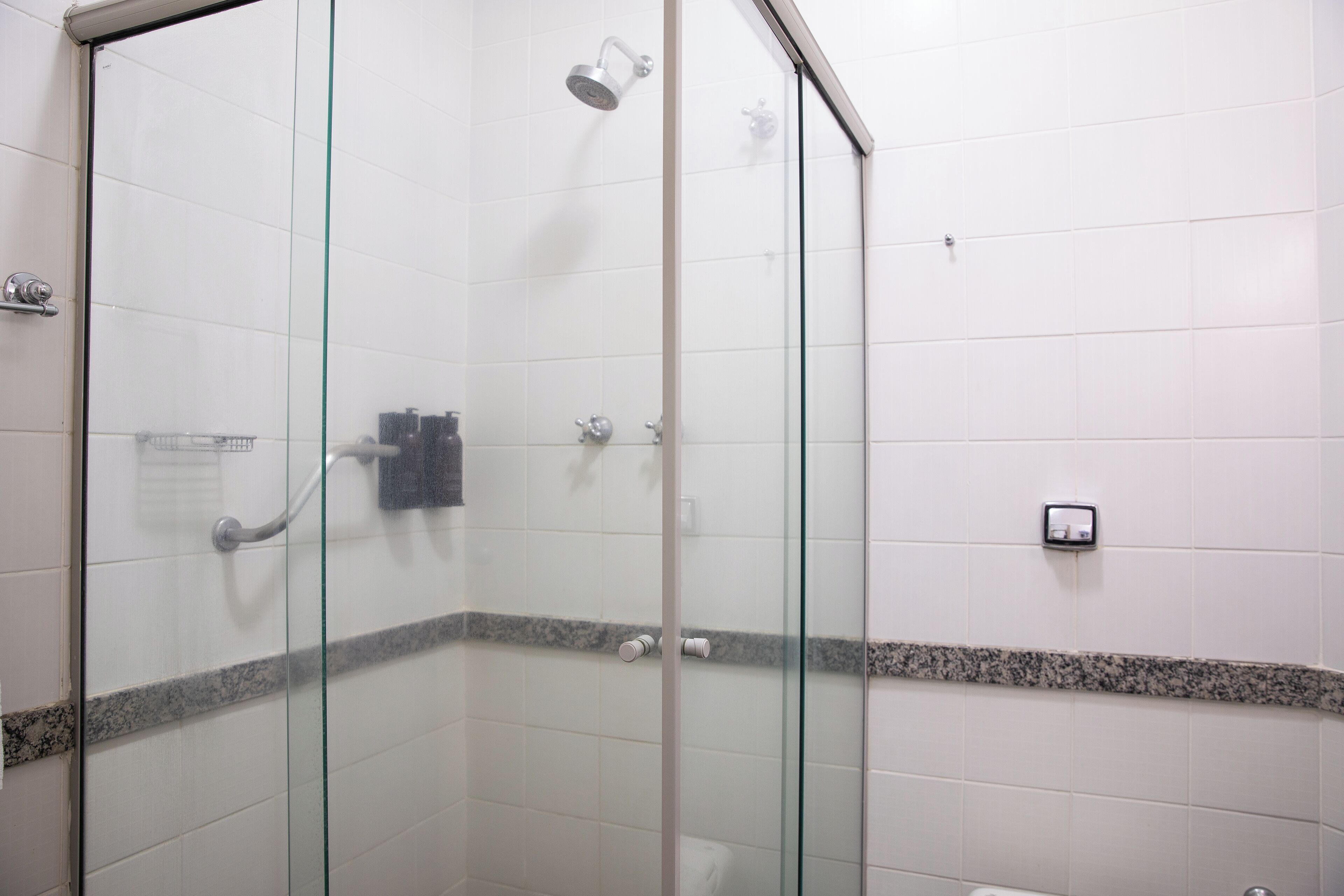 Un vestuario de ducha con paredes de vidrio, un cabezal de ducha manual, un perchero y un dispensador de jabón en la pared.